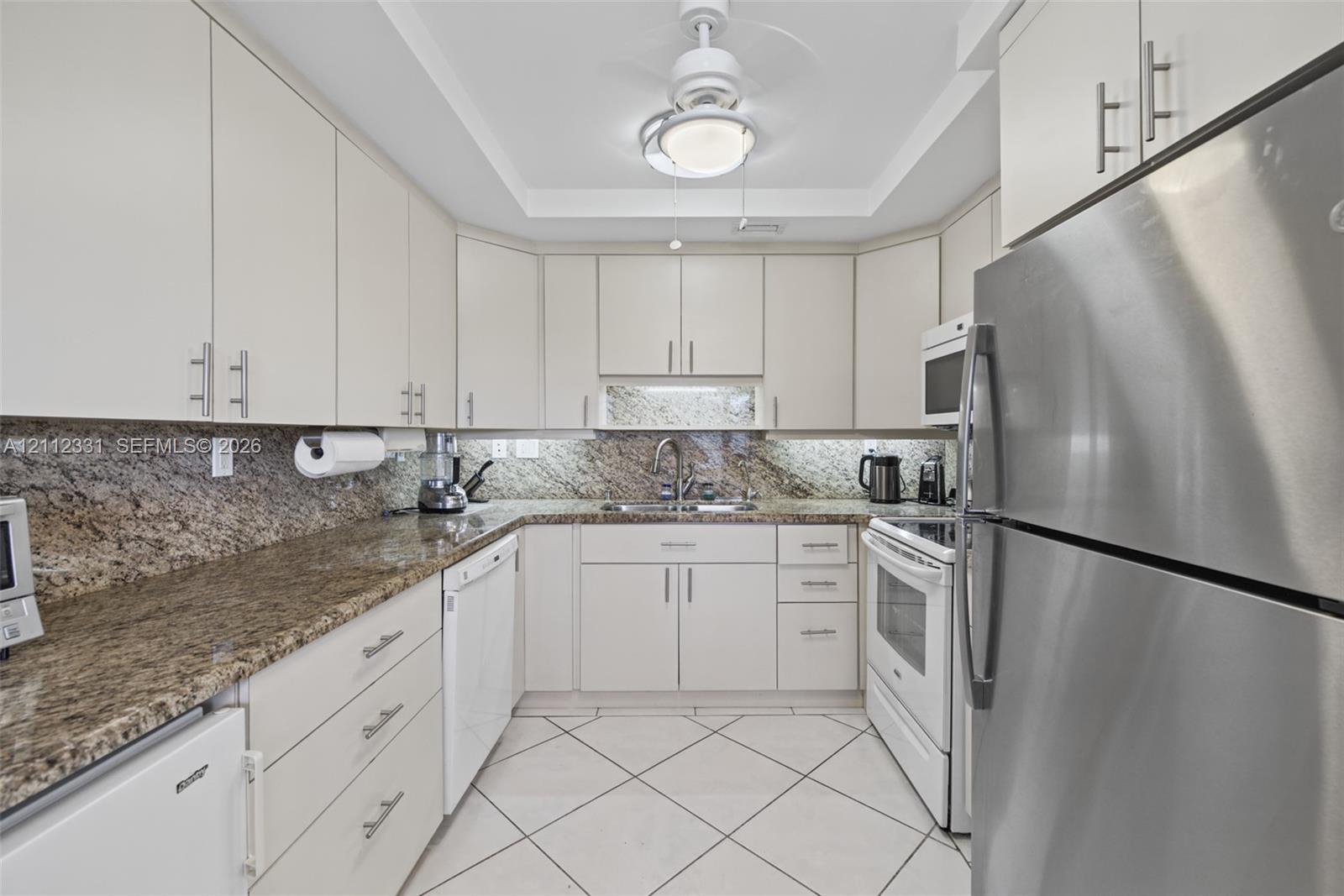 Kitchen featuring light-toned cabinetry with brushed metal pulls, stone-composite countertops, stainless steel refrigerator, white appliances, and diagonal-set tile flooring