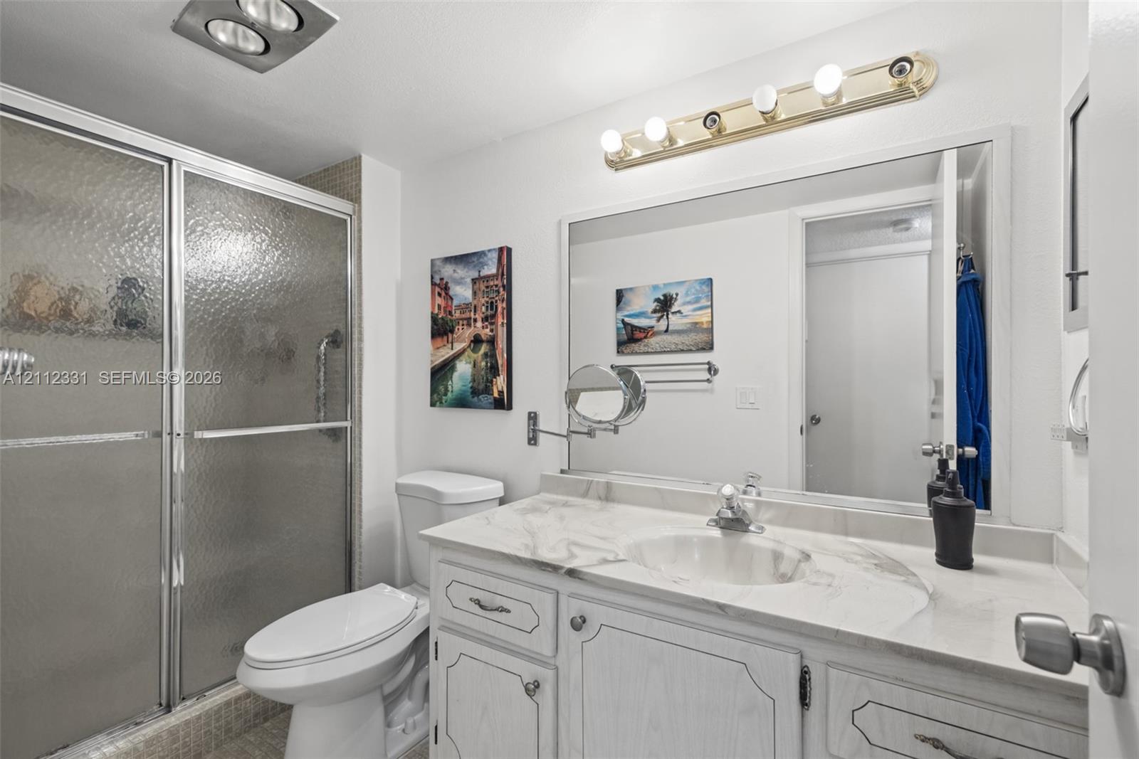 Bathroom featuring a vanity with a stone-top surface and integrated basin, a large wall-mounted mirror, a multi-bulb vanity light fixture, and a glass-enclosed shower with metal frame