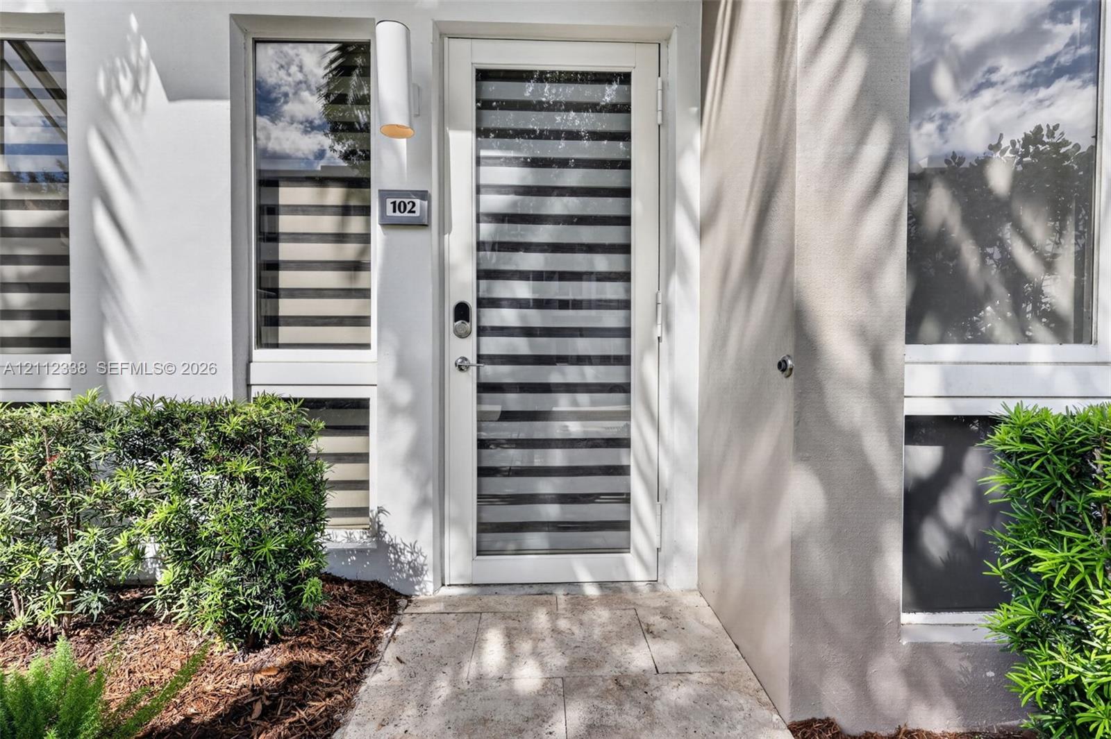 Exterior entry featuring a white door with horizontal glass panes, stone-finish paving, modern cylindrical wall-mounted lighting, and manicured shrubbery Exterior entry featuring a white door with horizontal glass panes, stone-finish paving, modern cylindrical wall-mounted lighting, and manicured shrubbery