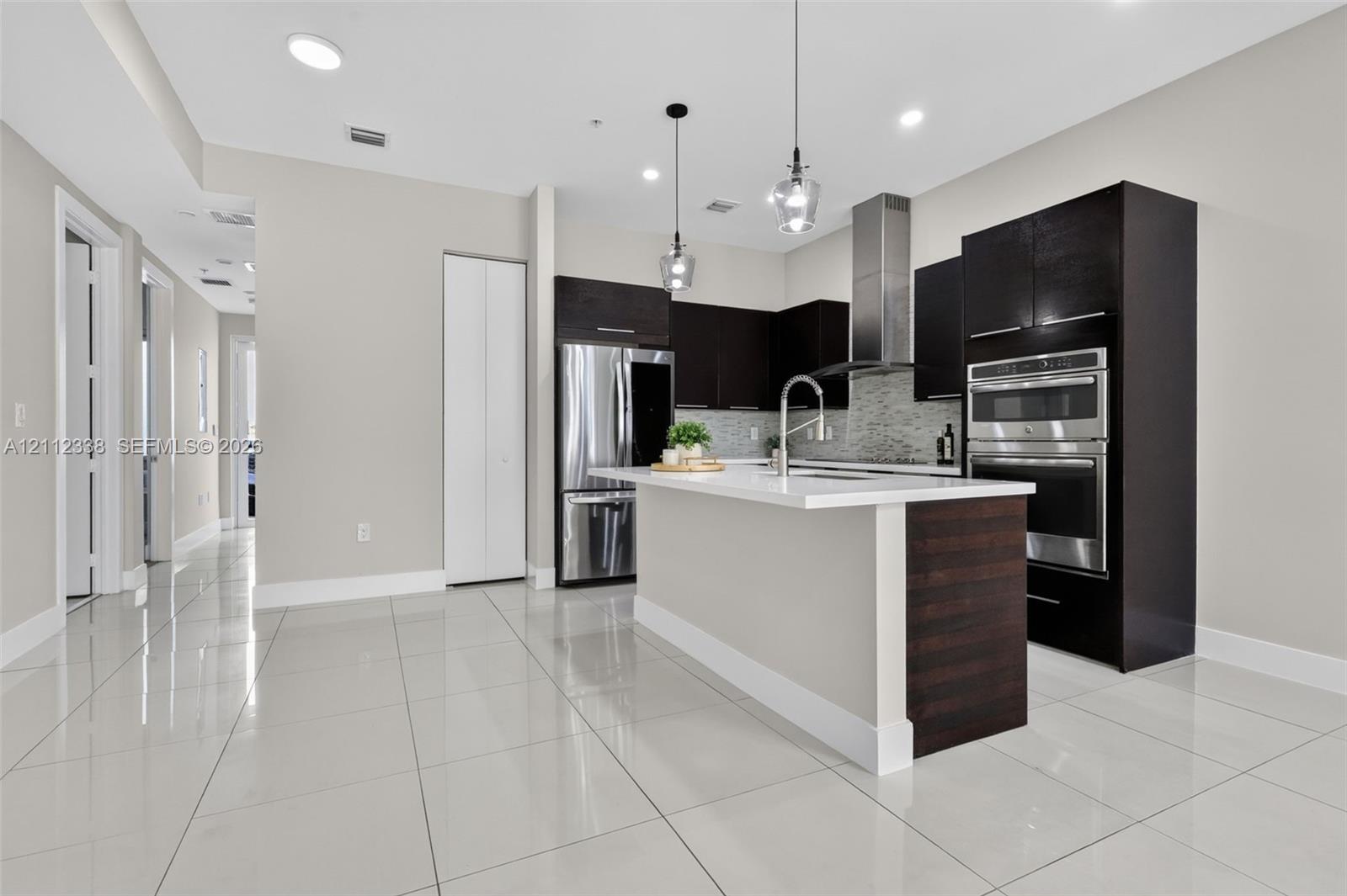 Kitchen featuring dark wood-finish cabinetry, a central island with white countertop, stainless steel double wall ovens, a stainless steel range hood, and tile flooring