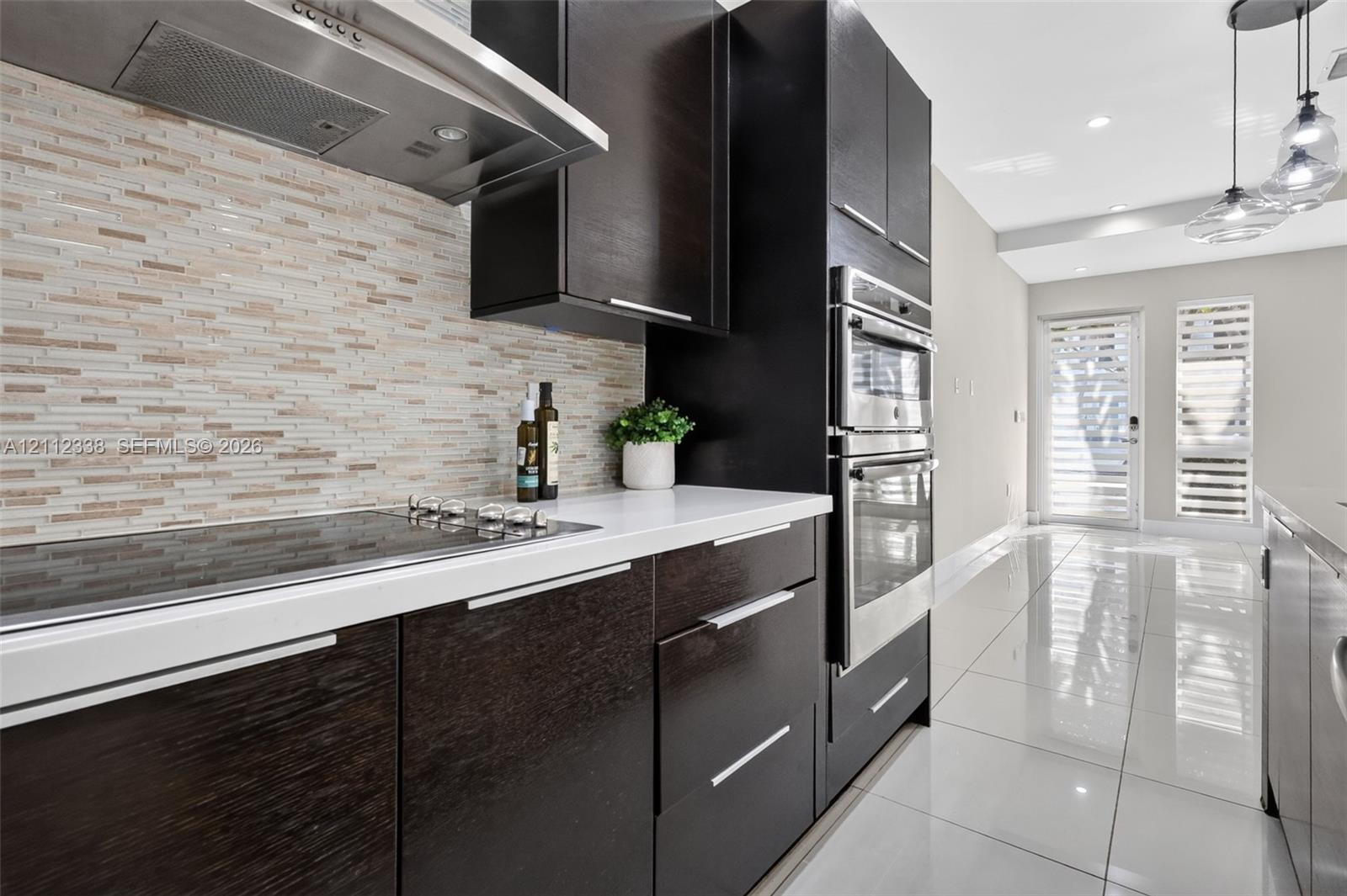 Kitchen featuring dark-finish flat-panel cabinetry, light-toned stone countertops, a linear mosaic tile backsplash, stainless steel built-in double ovens, and large-format porcelain tile flooring Kitchen featuring dark-finish flat-panel cabinetry, light-toned stone countertops, a linear mosaic tile backsplash, stainless steel built-in double ovens, and large-format porcelain tile flooring