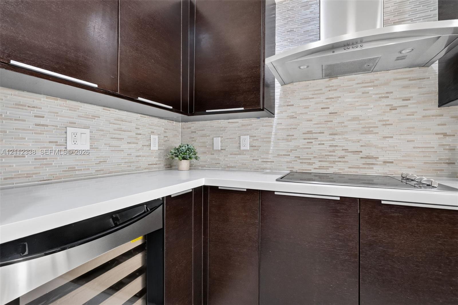 Dark wood-finish cabinetry featuring sleek hardware, white solid-surface countertops, horizontal linear tile backsplash, stainless steel range hood, and integrated under-counter beverage chiller Dark wood-finish cabinetry featuring sleek hardware, white solid-surface countertops, horizontal linear tile backsplash, stainless steel range hood, and integrated under-counter beverage chiller