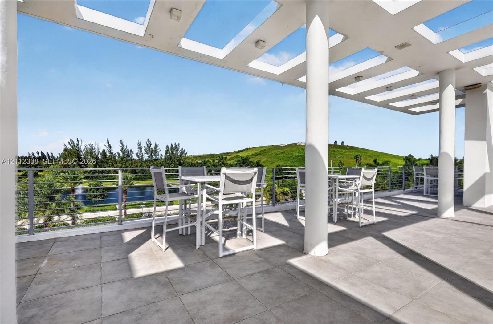 Covered terrace featuring a pergola-style ceiling, structural support columns, oversized tile flooring, and a metal cable railing system overlooking a landscape backdrop