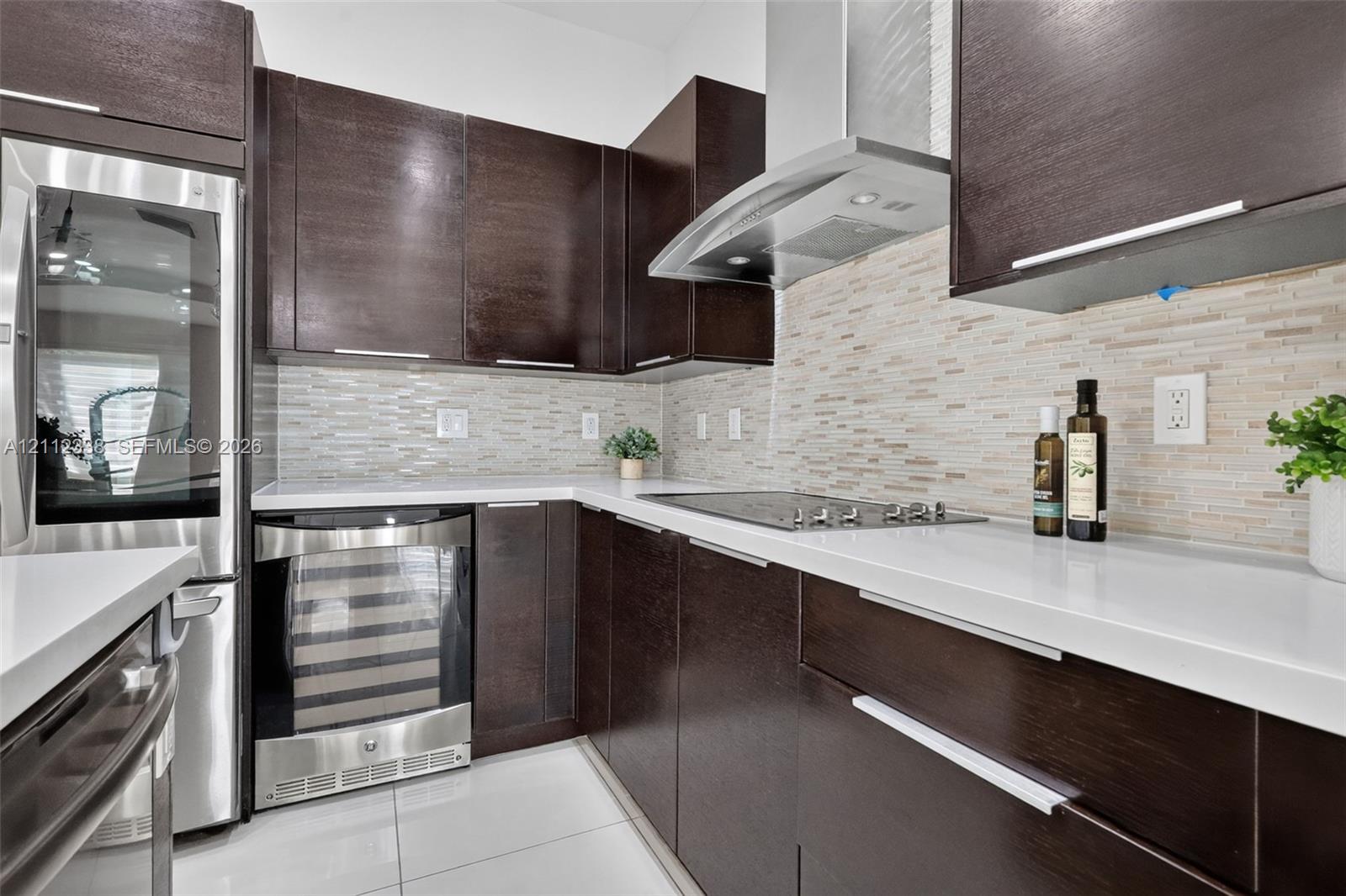 Dark wood cabinetry with modern hardware, white quartz countertops, stainless steel appliances, tiled backsplash, and light-tone tile flooring Dark wood cabinetry with modern hardware, white quartz countertops, stainless steel appliances, tiled backsplash, and light-tone tile flooring