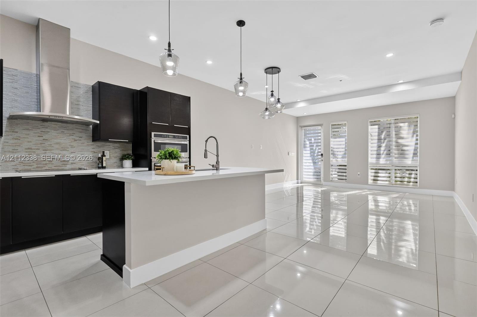 Kitchen featuring dark cabinetry, quartz countertops, a stone-tiled backsplash, stainless steel range hood, and polished tile flooring Kitchen featuring dark cabinetry, quartz countertops, a stone-tiled backsplash, stainless steel range hood, and polished tile flooring