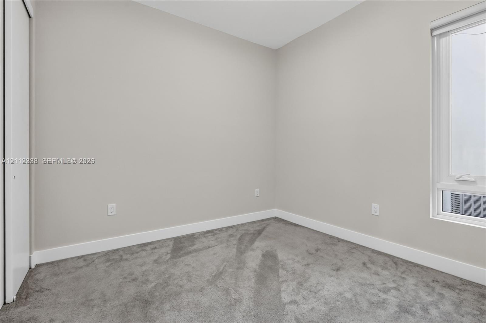 Neutral-toned interior space featuring soft wall-to-wall carpeting, crisp white walls, and a window with an integrated roller shade