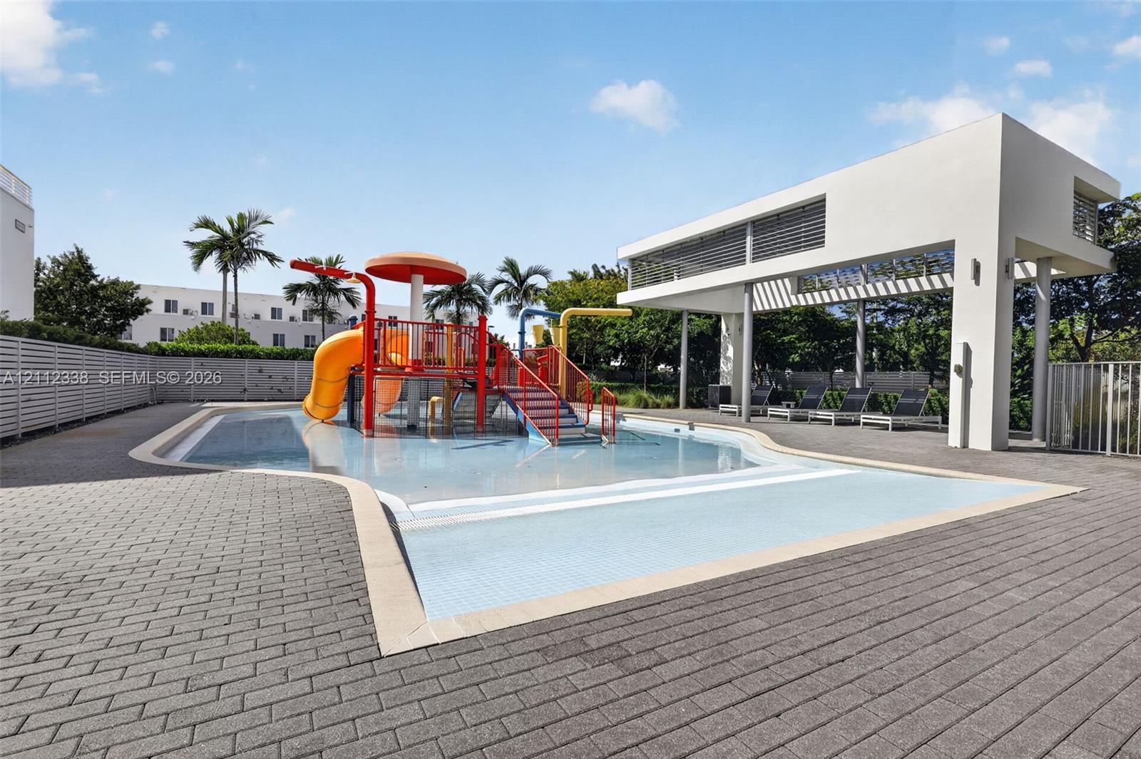 Splash pad area featuring integrated water slides, a central mushroom-shaped fountain, a concrete paver deck, and a modern open-air covered pavilion