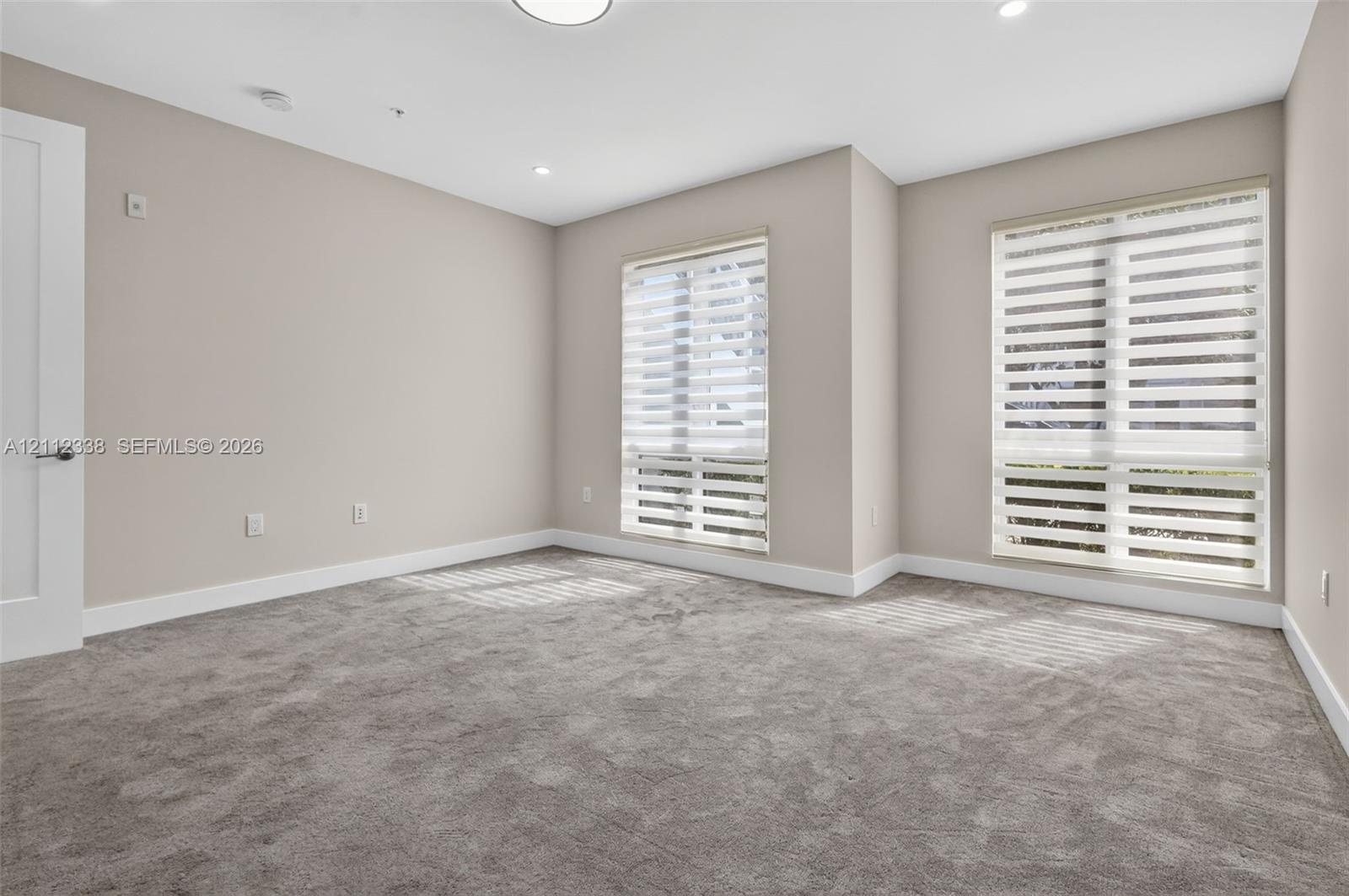 Neutral-toned interior featuring wall-to-wall carpeting, recessed ceiling lighting, and two windows equipped with dual-layer zebra shades