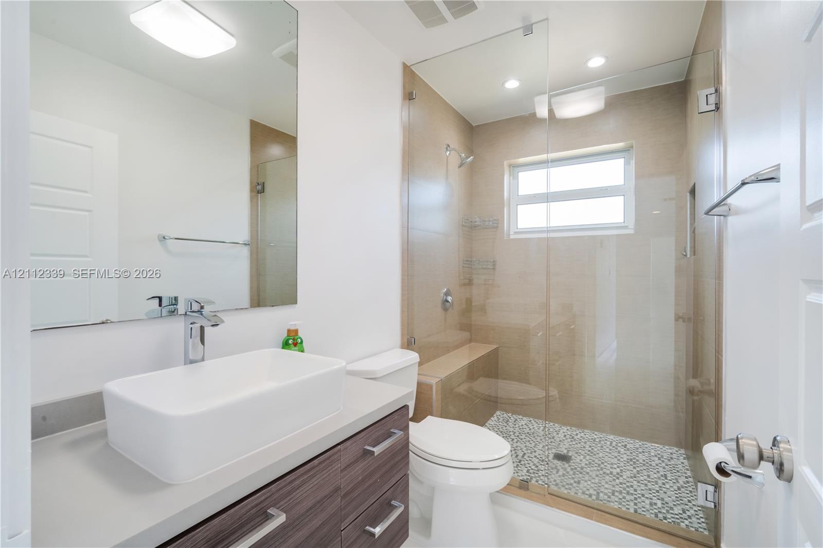 Bathroom featuring a vanity with a rectangular vessel sink, wood-finish cabinetry, a glass-enclosed shower with mosaic tile flooring, built-in shower seating, and a large wall-mounted mirror