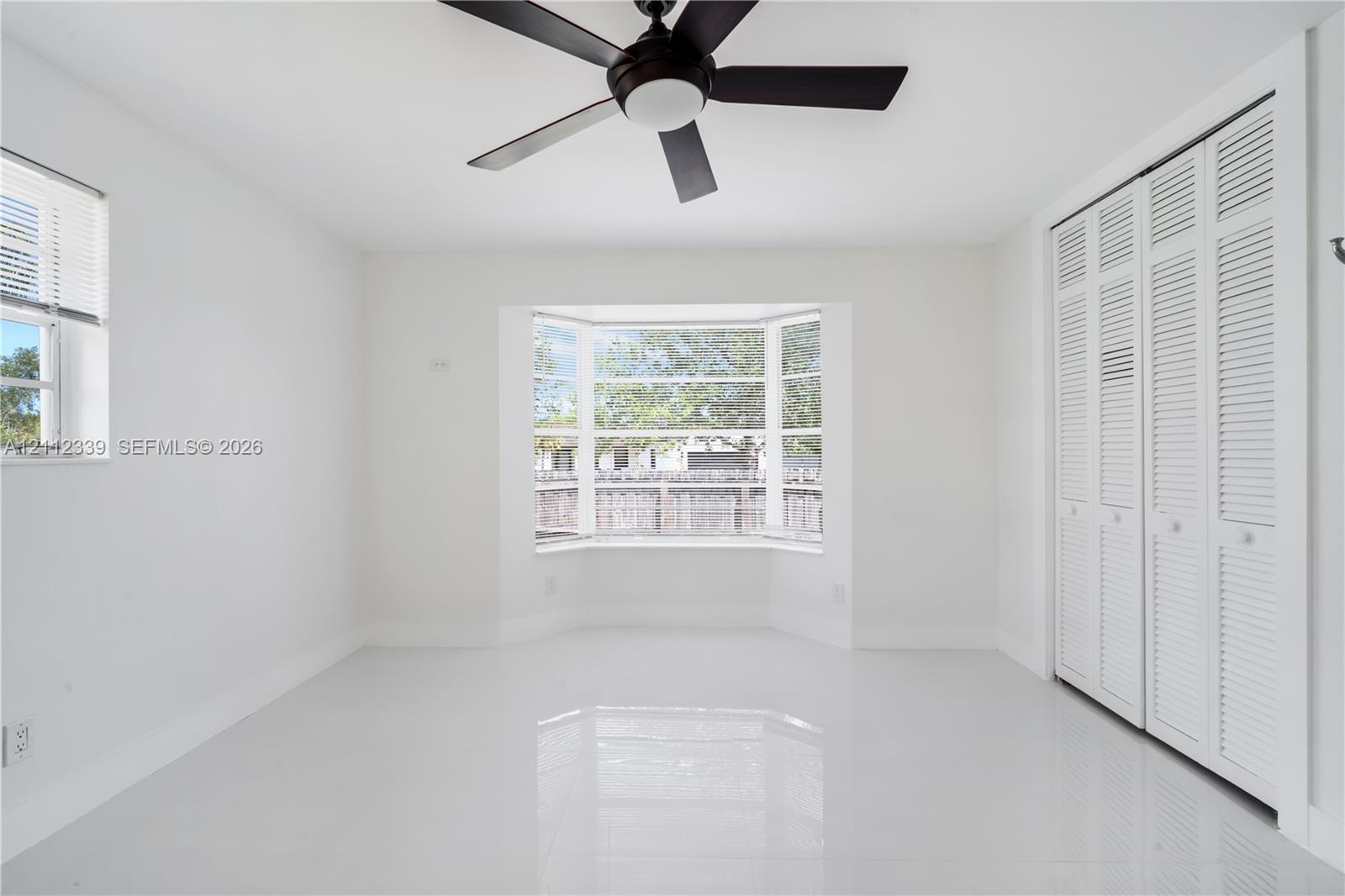 Room featuring large-format tile flooring, a multi-pane bay window, white louvered-style closet doors, and a ceiling-mounted fan with integrated lighting