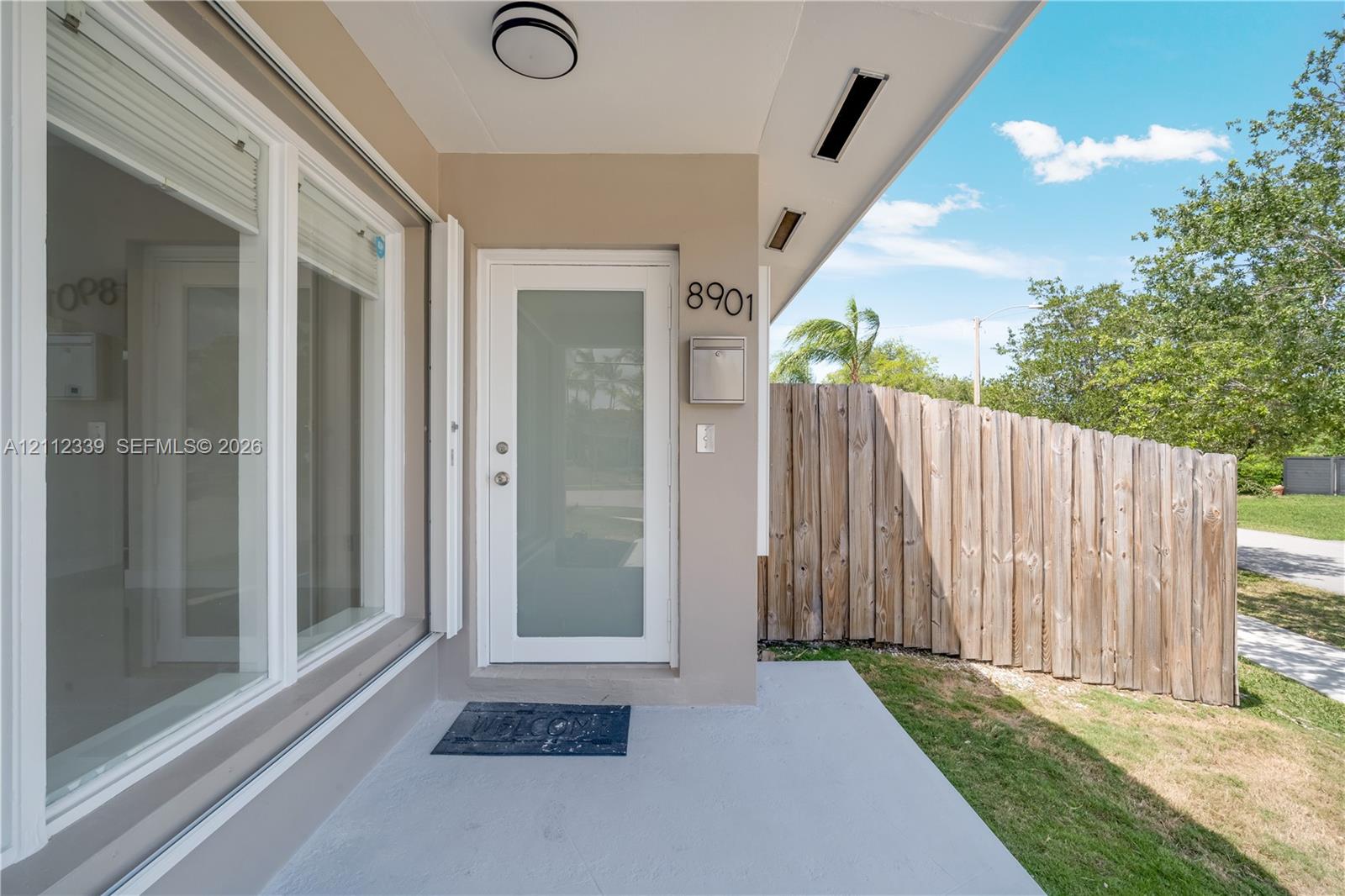Covered porch entry featuring a frosted glass panel front door, recessed lighting, exterior wall-mounted mailbox, and wood-slat privacy fencing