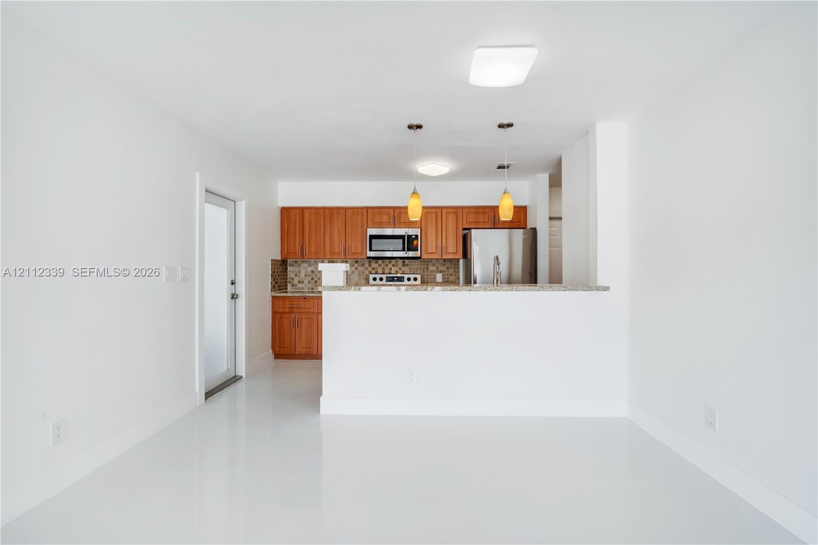 Renovated kitchen featuring wood cabinetry, granite countertops, a tiled backsplash, stainless steel appliances, and two hanging pendant lights