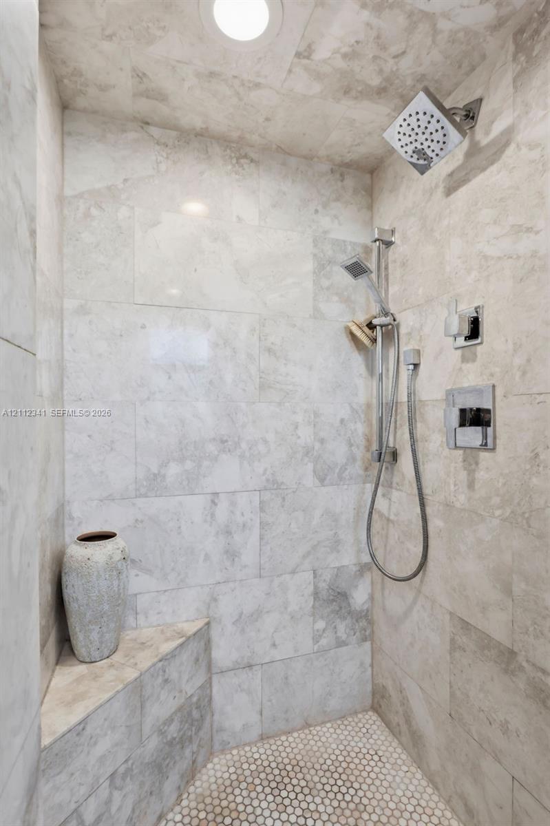 Walk-in shower featuring large-format stone tile wall cladding, hexagonal tile flooring, a built-in corner bench, a square rain-style showerhead, and a multi-function handheld shower attachment with polished chrome hardware