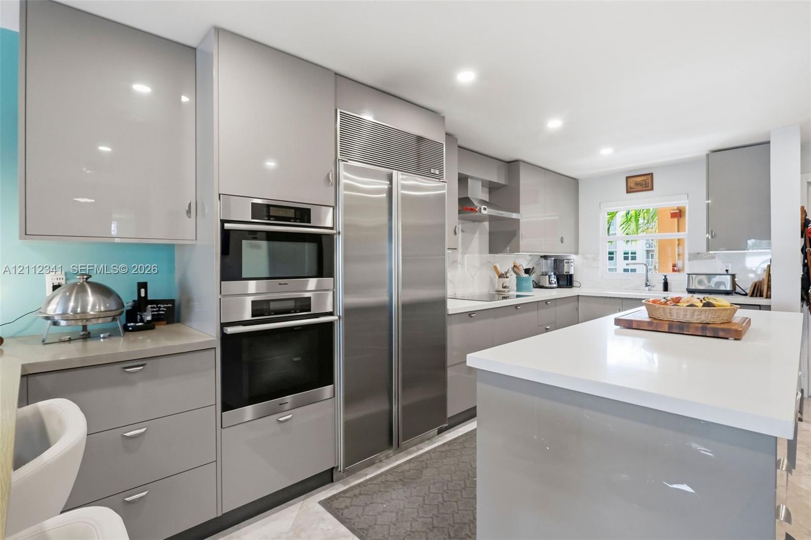 Modern kitchen featuring high-gloss cabinetry, stainless steel professional-grade refrigerator, integrated dual wall ovens, solid-surface countertops, and a central island with waterfall edge