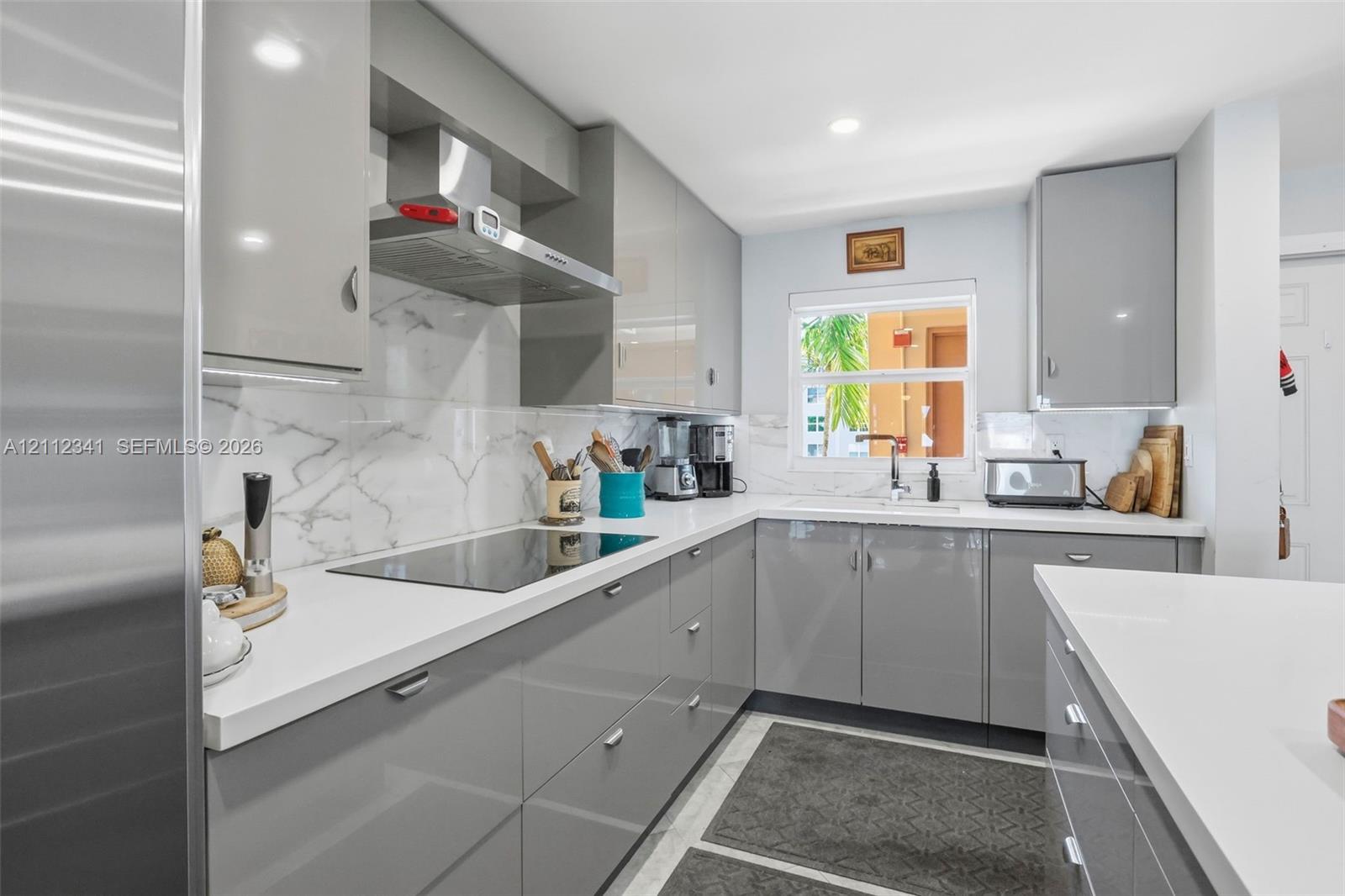 Kitchen featuring high-gloss gray slab cabinetry, solid-surface countertops, full-height stone-patterned backsplash, stainless steel range hood, and integrated cooktop