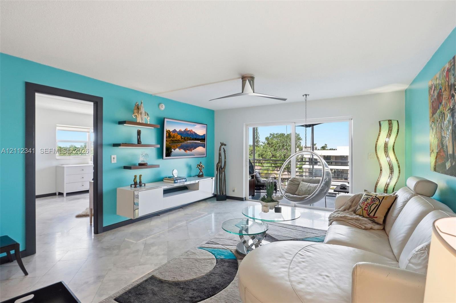 Living area featuring stone-finish flooring, a contemporary ceiling fan, built-in floating cabinetry with illuminated shelving, and sliding glass doors opening to a balcony Living area featuring stone-finish flooring, a contemporary ceiling fan, built-in floating cabinetry with illuminated shelving, and sliding glass doors opening to a balcony