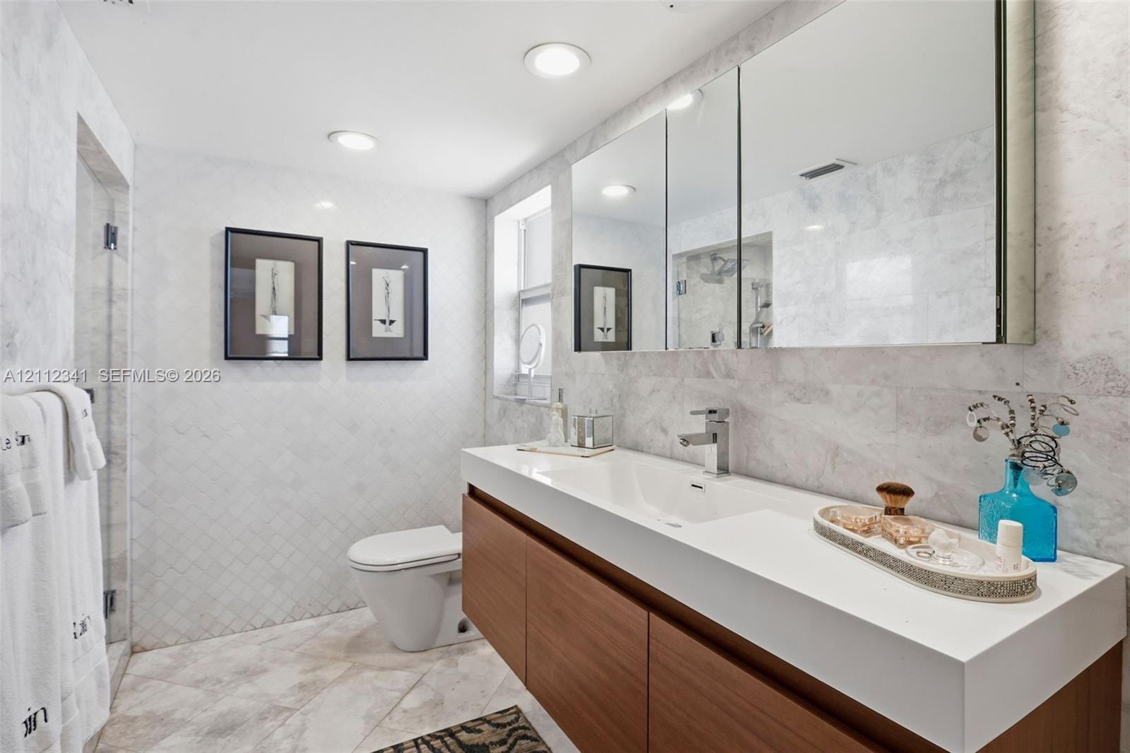 Modern floating vanity with a white countertop and rectangular sink, wood-finish cabinetry, recessed ceiling lighting, large triple-panel medicine cabinet, and full-wall stone tile surround