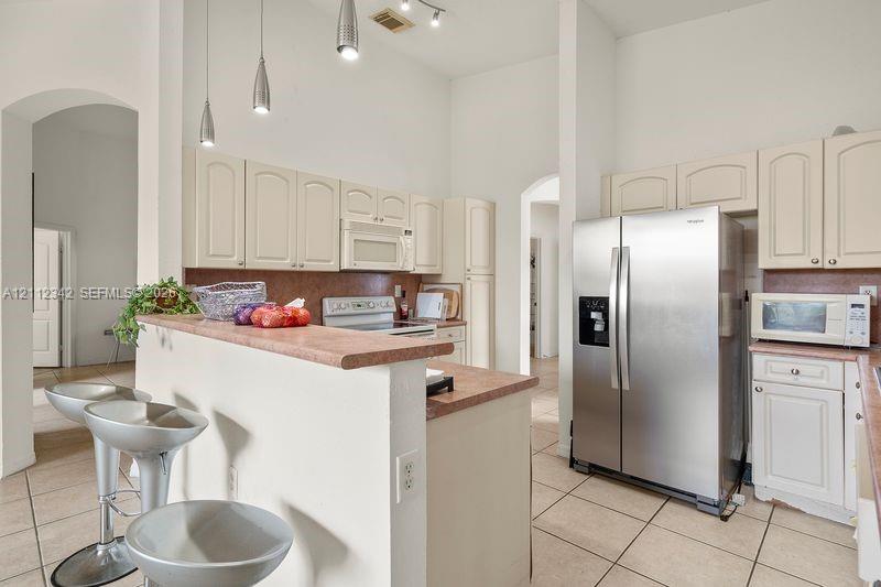 Kitchen featuring high ceilings, off-white cabinetry, solid-surface countertops with a breakfast bar, ceramic tile flooring, and stainless steel side-by-side refrigerator