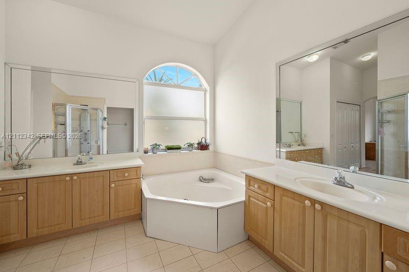 Bathroom featuring dual wood-finish vanities with solid-surface countertops, a corner soaking tub, an arched window, and dual wall-mounted vanity mirrors