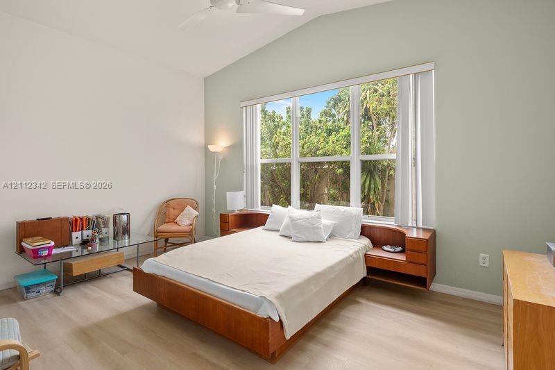 Vaulted ceiling with integrated paddle fan, expansive windows, wood-finish flooring, and built-in bedside cabinetry