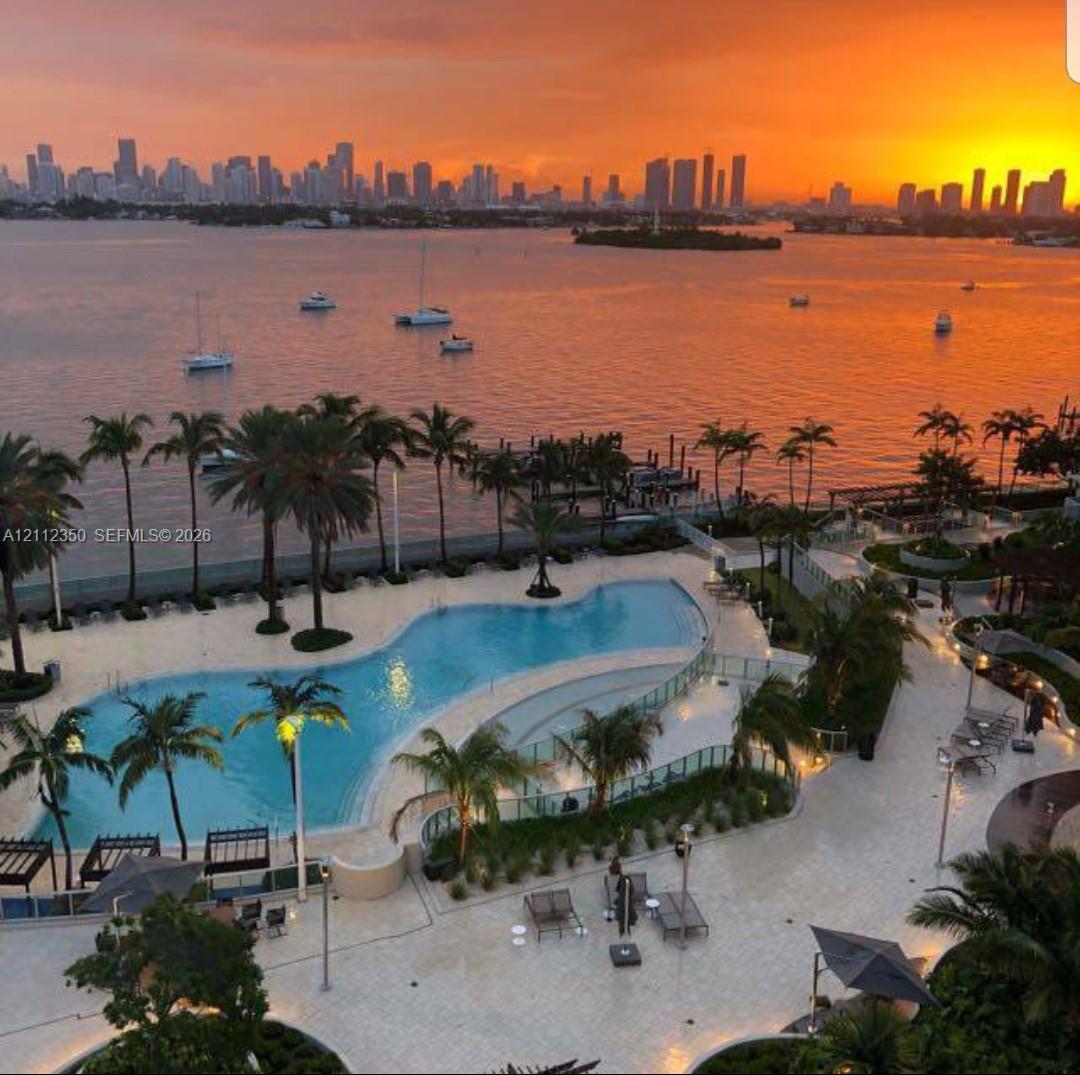 Waterfront swimming pool with sun shelf, expansive paved deck, integrated tropical landscaping, and panoramic skyline horizon