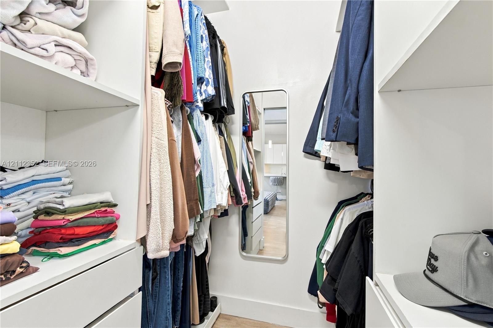 Walk-in closet featuring white custom built-in shelving, hanging rods, integrated drawer storage, and a wall-mounted rectangular mirror