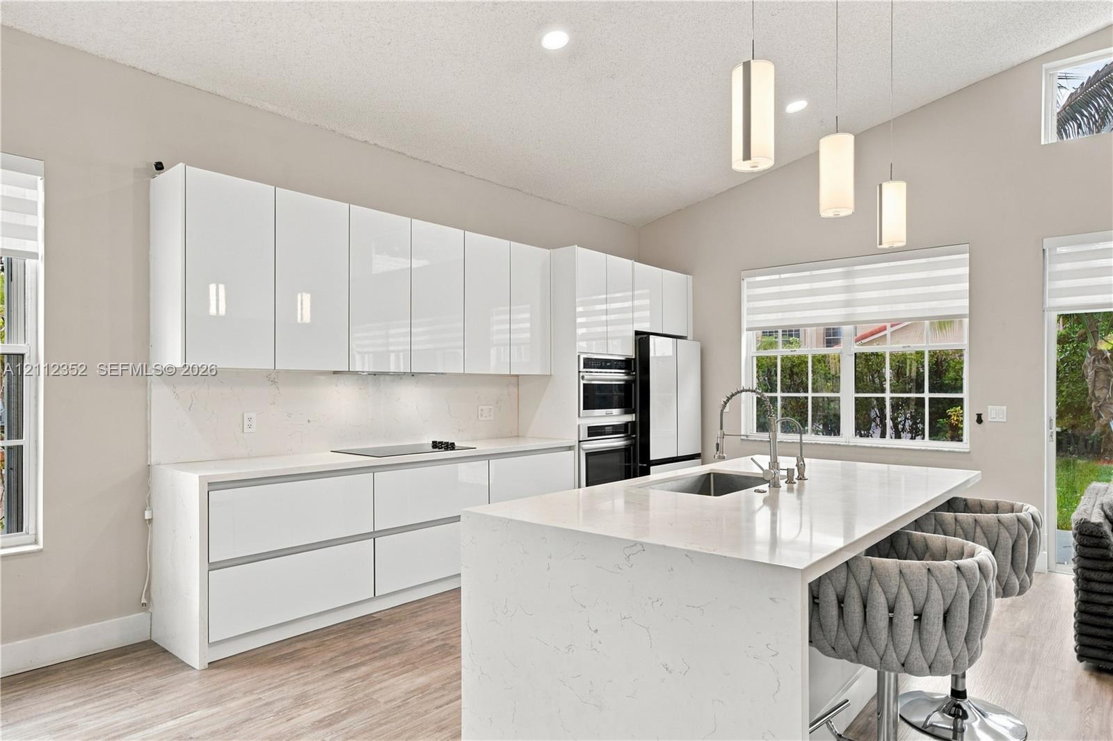 Modern kitchen featuring flat-panel white cabinetry, stone-surface countertops, a waterfall-edge center island with an undermount sink, built-in double wall ovens, and pendant lighting