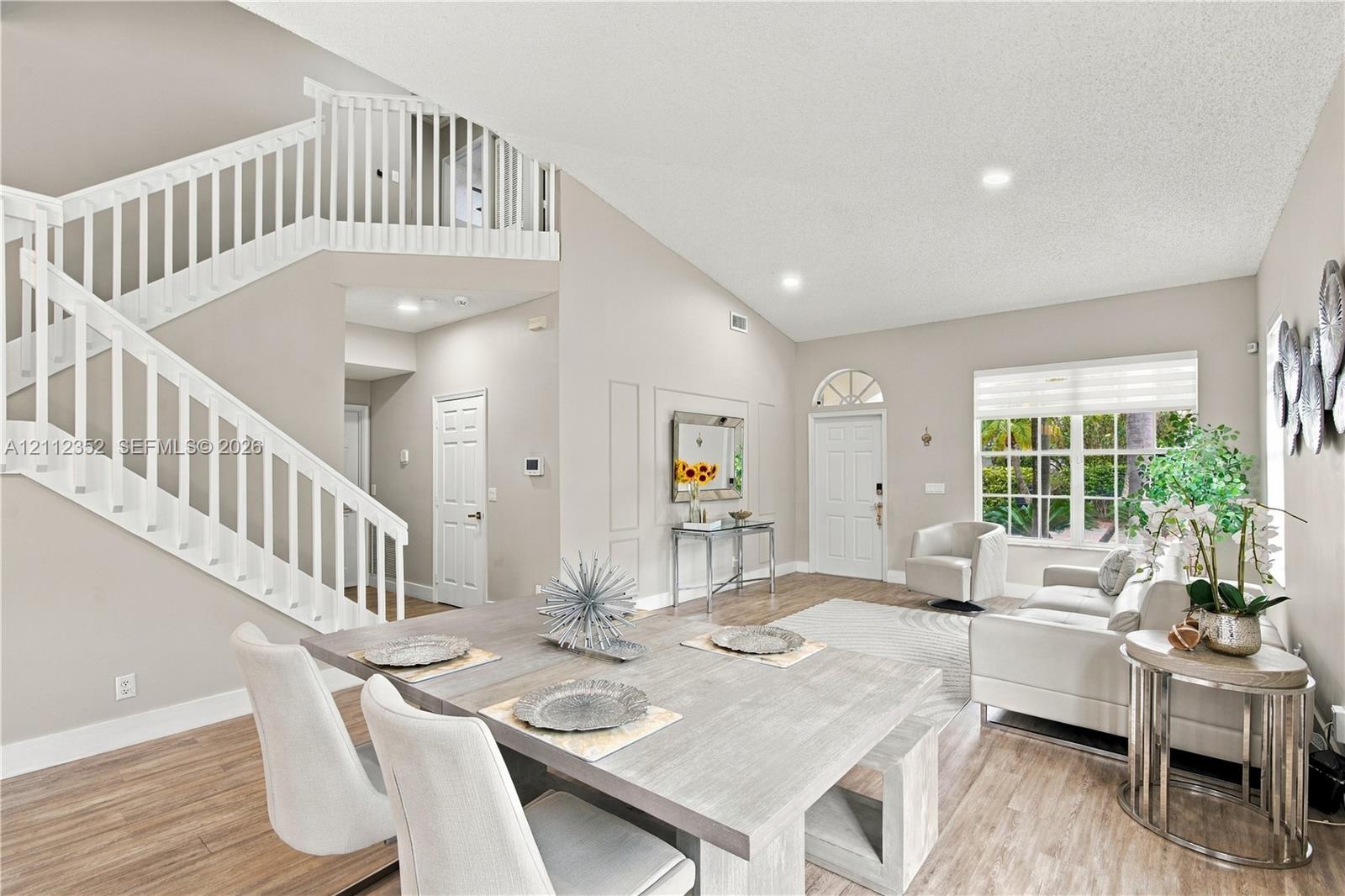 Open-concept interior featuring vaulted ceilings, wood-finish flooring, a white staircase with decorative balustrade, and multi-paned windows with a transom Open-concept interior featuring vaulted ceilings, wood-finish flooring, a white staircase with decorative balustrade, and multi-paned windows with a transom