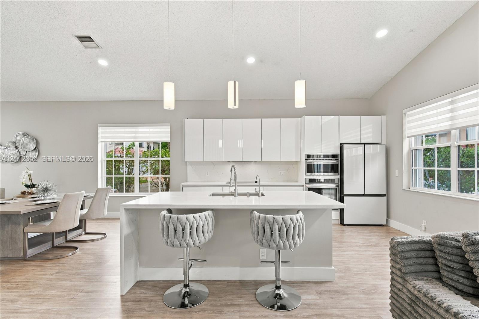 Modern kitchen featuring gloss-finish cabinetry, a white countertop island with an undermount sink, three pendant lights, built-in stainless steel appliances, and wood-finish flooring