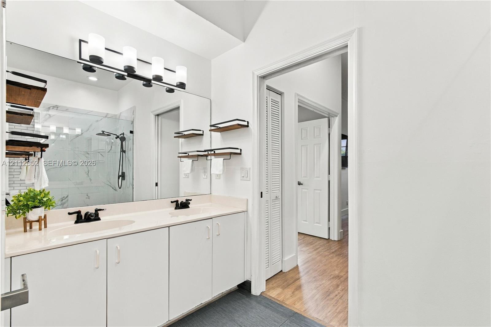 Dual-sink vanity with solid-surface countertop, full-width wall mirror, four-light vanity fixture, matte black hardware, and shower featuring large-format wall tile