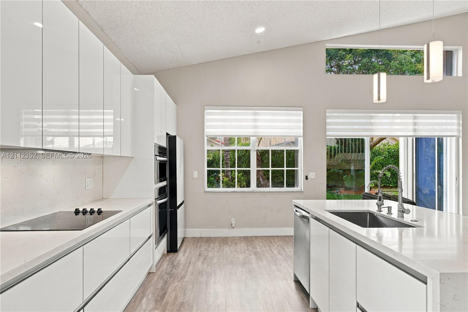 Modern kitchen featuring high-gloss white cabinetry, quartz countertops, a central island with a stainless steel sink, wood-finish flooring, and vaulted ceilings with clerestory windows