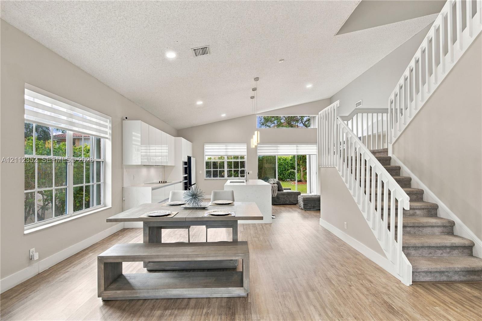 Open-concept interior featuring wood-finish flooring, vaulted ceilings, high-gloss white cabinetry, a center kitchen island with waterfall-edge countertop, and a staircase with white balusters Open-concept interior featuring wood-finish flooring, vaulted ceilings, high-gloss white cabinetry, a center kitchen island with waterfall-edge countertop, and a staircase with white balusters