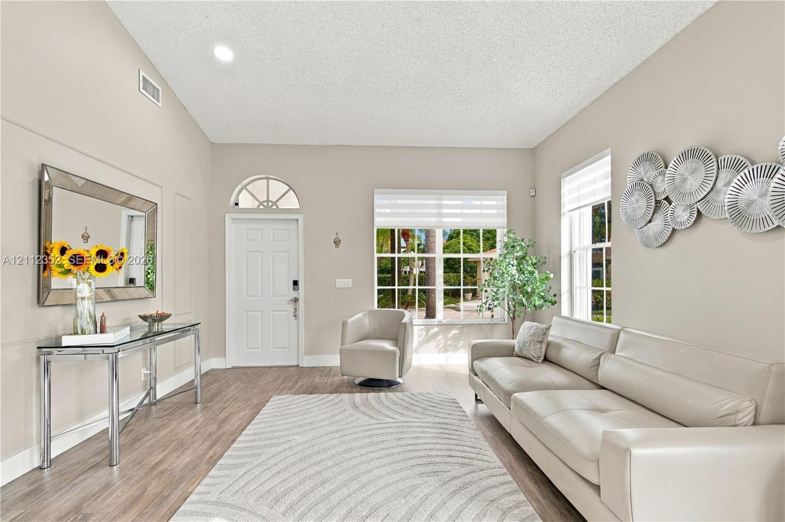 High-ceilinged room featuring wood-finish flooring, a multi-pane entry door with a semi-circular transom window, and oversized windows with integrated blinds High-ceilinged room featuring wood-finish flooring, a multi-pane entry door with a semi-circular transom window, and oversized windows with integrated blinds