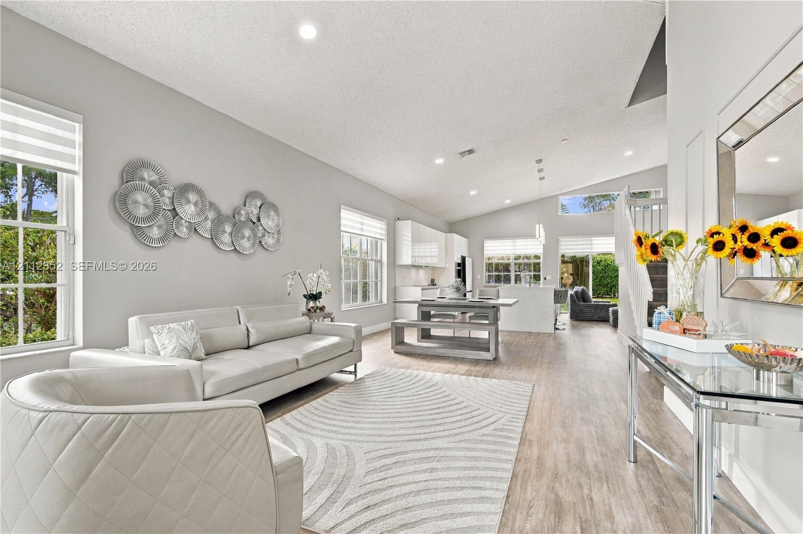 Open-concept interior featuring vaulted ceilings, wood-finish flooring, recessed lighting, and multiple windows with integrated blinds Open-concept interior featuring vaulted ceilings, wood-finish flooring, recessed lighting, and multiple windows with integrated blinds