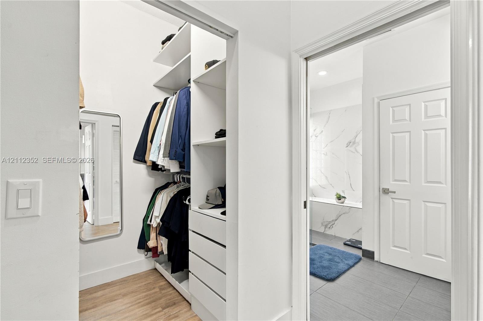 Walk-in closet featuring custom white shelving, integrated drawers, and wood-finish flooring, accompanied by a bathroom showcasing a stone-tile shower surround and large-format floor tile