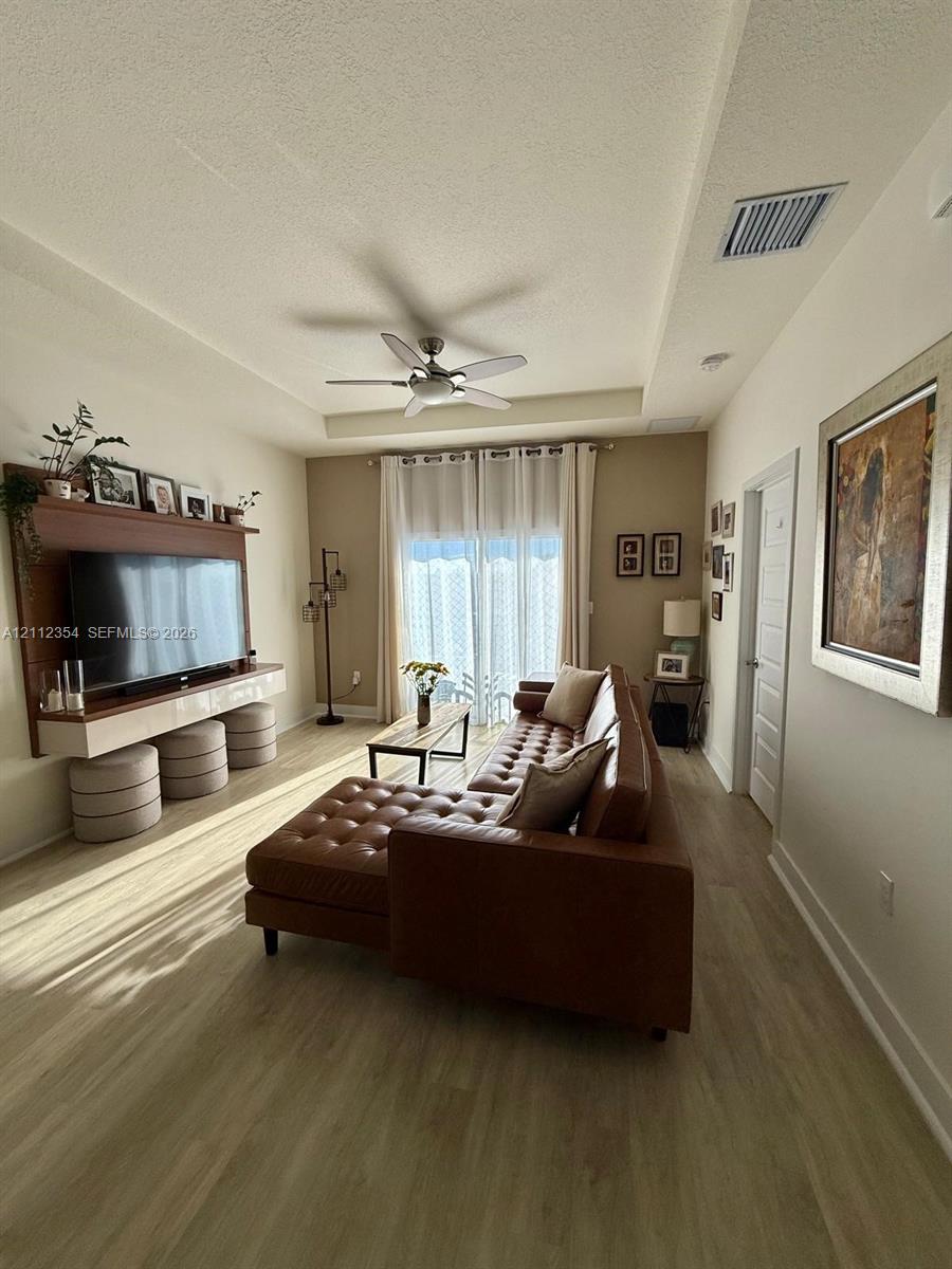 Tray ceiling with a central multi-blade ceiling fan, wood-finish flooring, and a wall-mounted built-in media console with integrated shelving