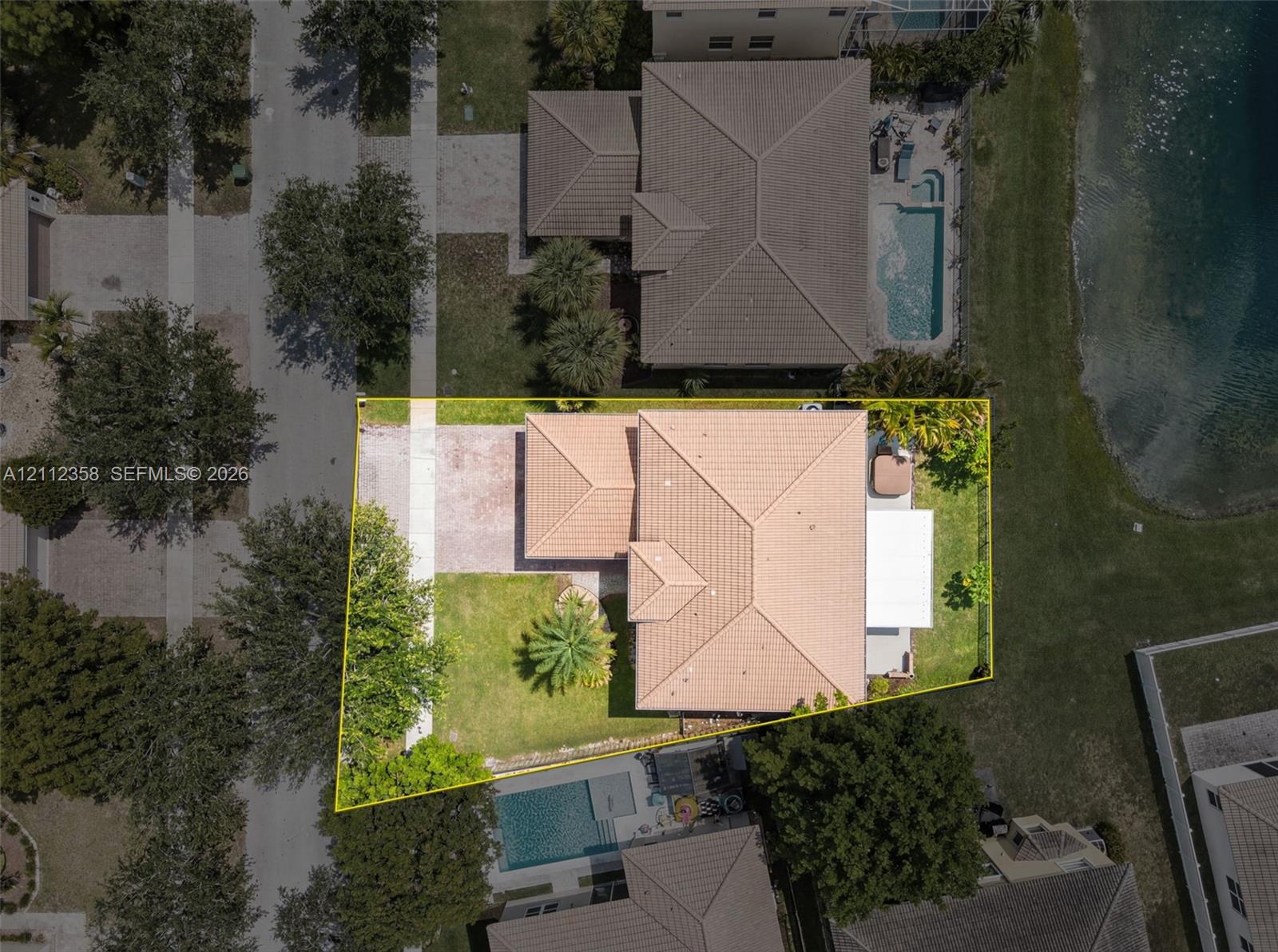 Aerial view of property featuring a tile roof, paved driveway, landscaped lawn with mature trees, and a private backyard patio