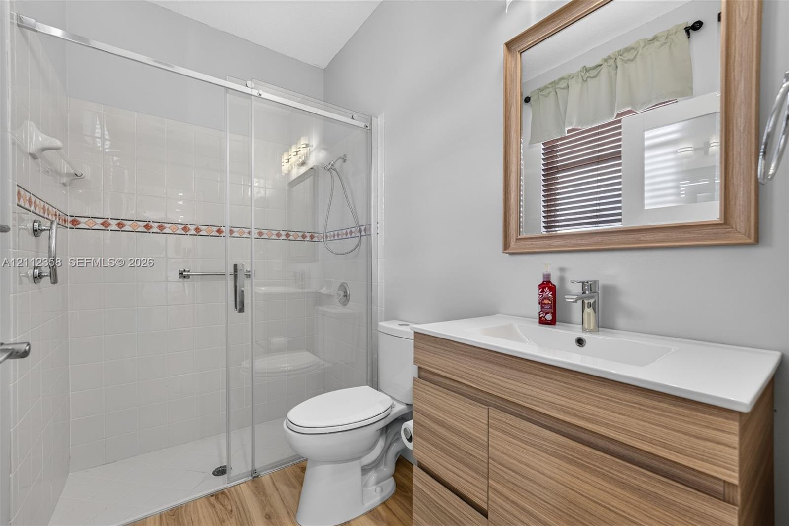Bathroom featuring a wood-finish vanity with a rectangular sink, walk-in glass-enclosed shower with tile surround, wood-finish flooring, and a framed wall mirror