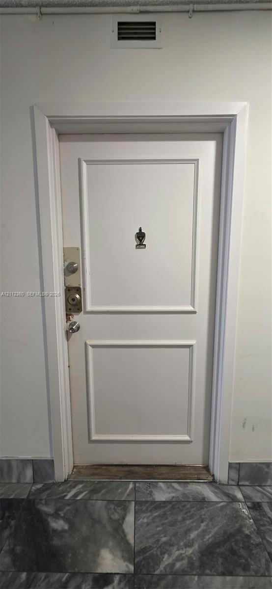 White paneled door featuring a decorative metal door knocker, dual deadbolt locks, and a polished metal door handle, set against stone-finish flooring