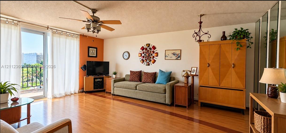 Living area featuring wood-finish flooring, a ceiling fan with integrated lighting, sliding glass door access to a balcony, and mirrored closet doors