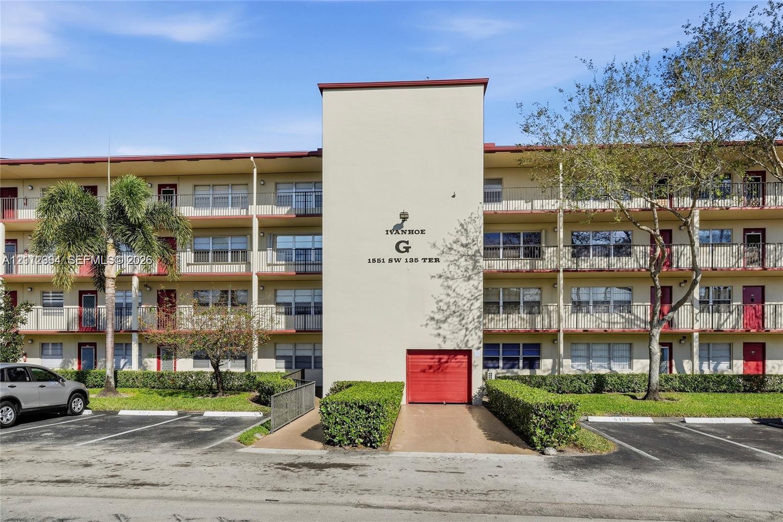 Multi-story residential building exterior featuring a central stucco facade, horizontal balconies with metal railings, red-accented doors, and asphalt parking area Multi-story residential building exterior featuring a central stucco facade, horizontal balconies with metal railings, red-accented doors, and asphalt parking area