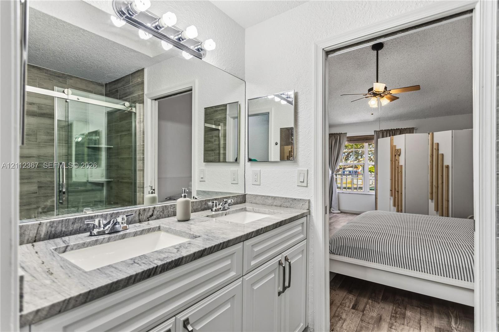 Bathroom featuring a dual-sink vanity with stone countertop, oversized wall-to-wall mirror, glass-enclosed shower with tiled surround, and integrated cabinetry