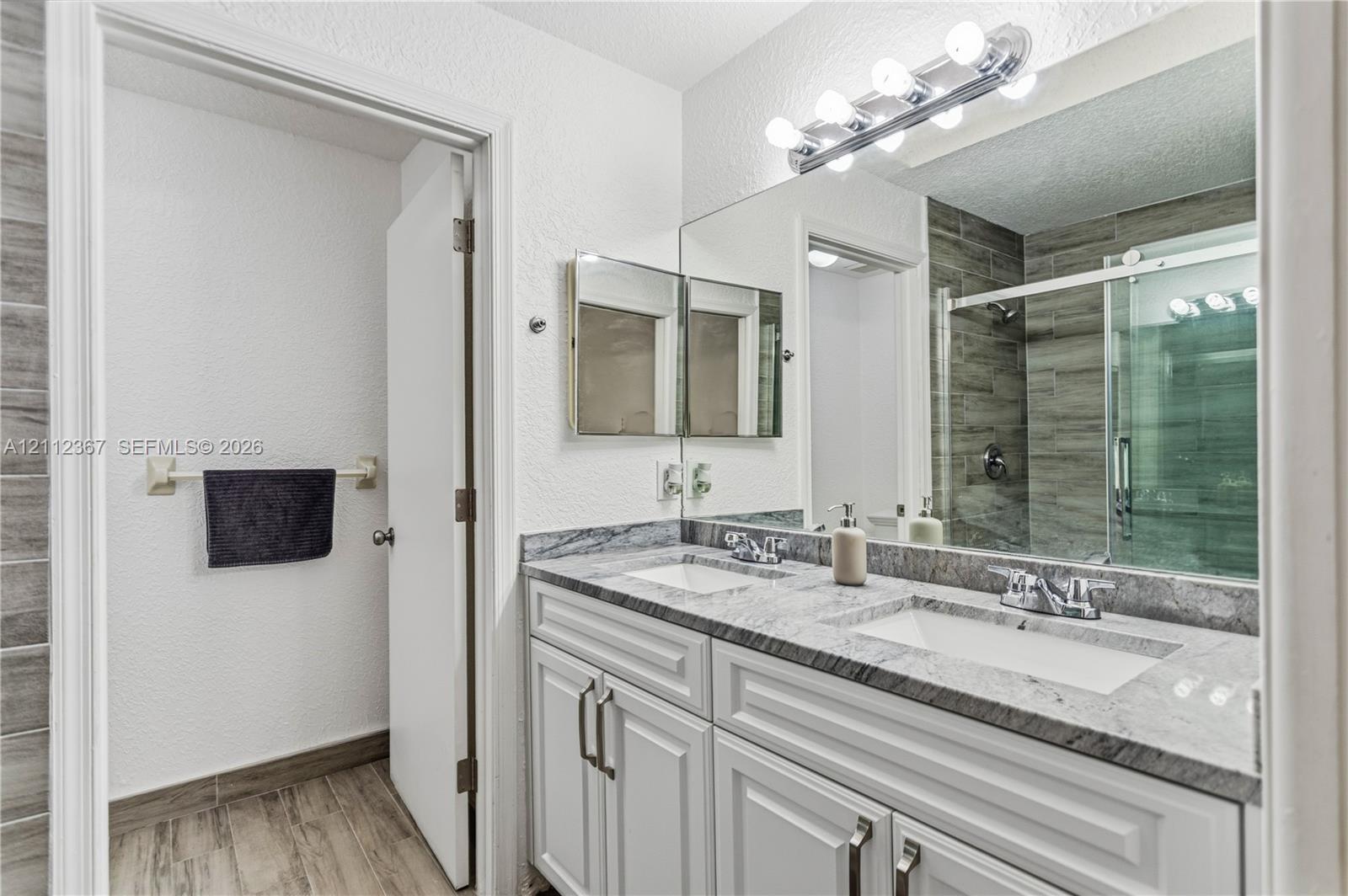 Dual-sink vanity with stone-finish countertop, white shaker-style cabinetry, wood-finish flooring, full-width wall mirror, and shower enclosure with horizontal tile surround
