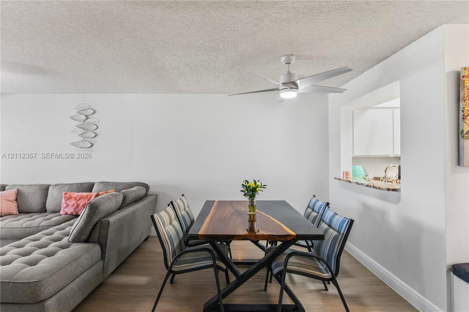 Neutral interior space featuring wood-finish flooring, a contemporary white ceiling fan with integrated lighting, and a kitchen pass-through window with a stone-surface counter