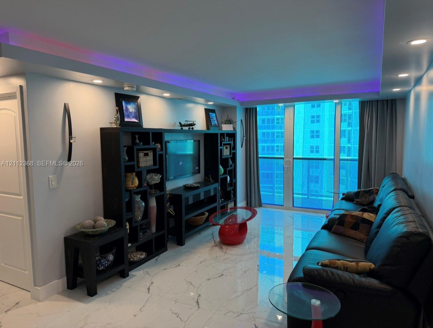 Large-format stone-finish floor tiling, recessed ceiling lighting with integrated colored accent lighting, built-in shelving unit, and floor-to-ceiling glass paneling leading to a balcony