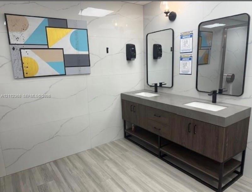 Double vanity featuring a thick concrete-style countertop with integrated sinks, wood-finish base cabinetry, large wall-mounted rectangular mirrors with matte black frames, and full-height veined tile wall surfacing