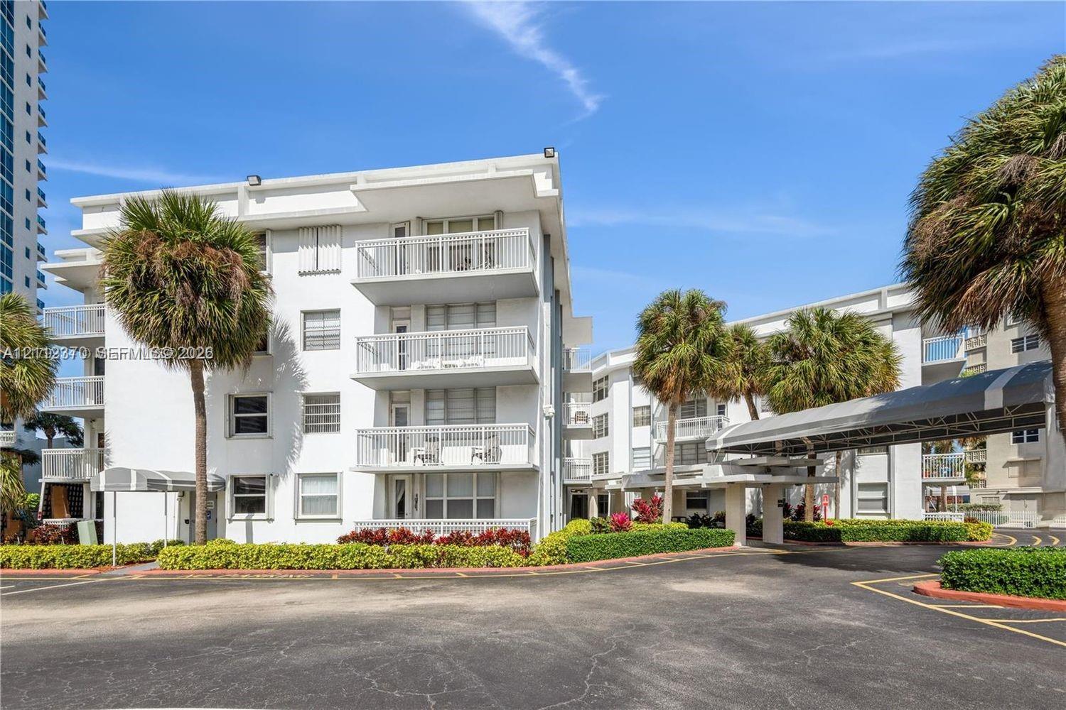 Mid-rise building exterior featuring private balconies with white railings, a covered porte-cochre entry, and mature palm tree landscaping Mid-rise building exterior featuring private balconies with white railings, a covered porte-cochre entry, and mature palm tree landscaping