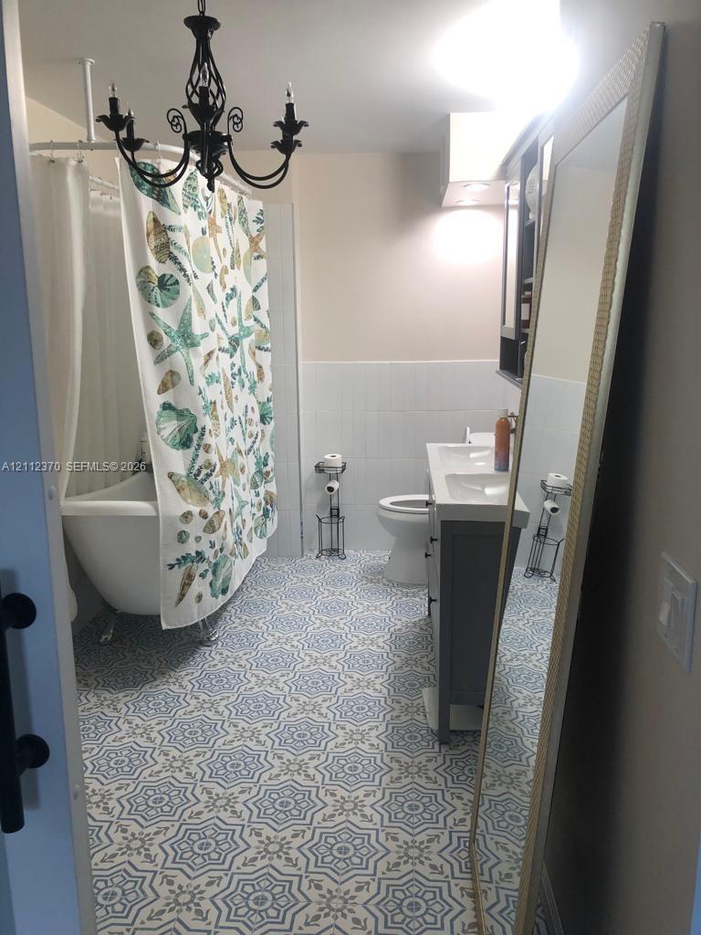 Clawfoot tub, patterned tile flooring, dual-basin vanity with gray cabinetry, and decorative wrought-iron chandelier