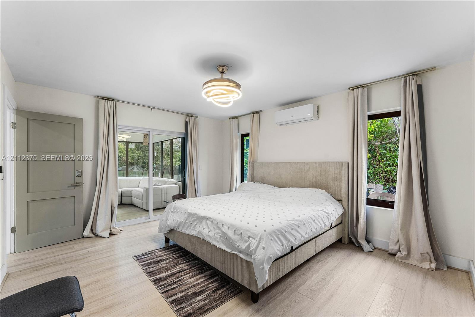 Light-toned wood-finish flooring, modern circular tiered ceiling light fixture, wall-mounted ductless air conditioning unit, sliding glass doors, and a solid-panel interior door
