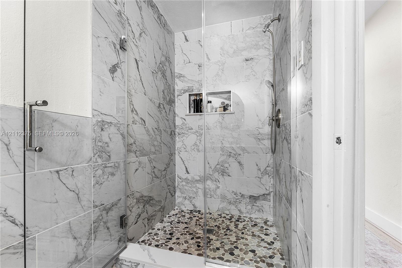 Walk-in shower featuring floor-to-ceiling stone-pattern tile, a glass enclosure, a pebble-finish floor, a recessed wall niche, and a handheld showerhead fixture